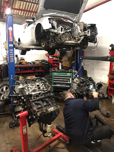 An EE Motors technician working on an engine, with a car raised on the ramp behind